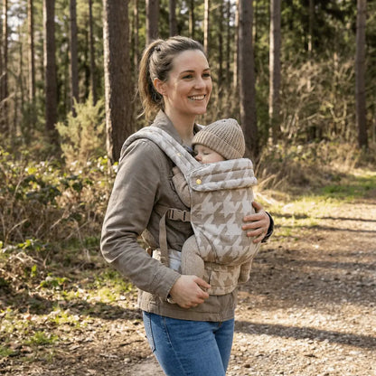 Needella Embrace Babytrage Im Hahnentrittmuster in Beige, Mutter trägt ihr Baby nah am Körper im Alltag