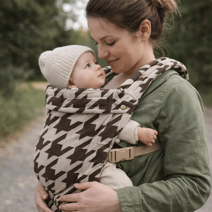Needella Embrace Babytrage im Hahnentrittmuster in Kaffee, Mutter trägt ihr Baby nah am Körper im Alltag