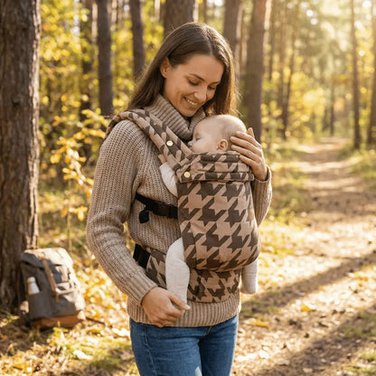Needella Embrace Babytrage im Hahnentrittmuster in Karamell, Mutter trägt ihr Baby nah am Körper im Alltag