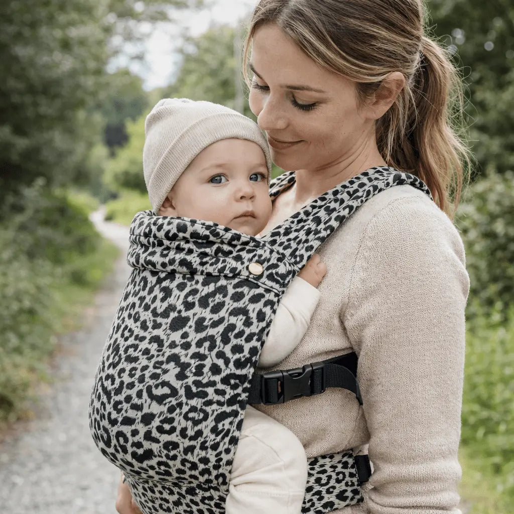 Needella Embrace Babytrage im Leopardmuster in Weiß, Mutter trägt ihr Baby nah am Körper im Alltag